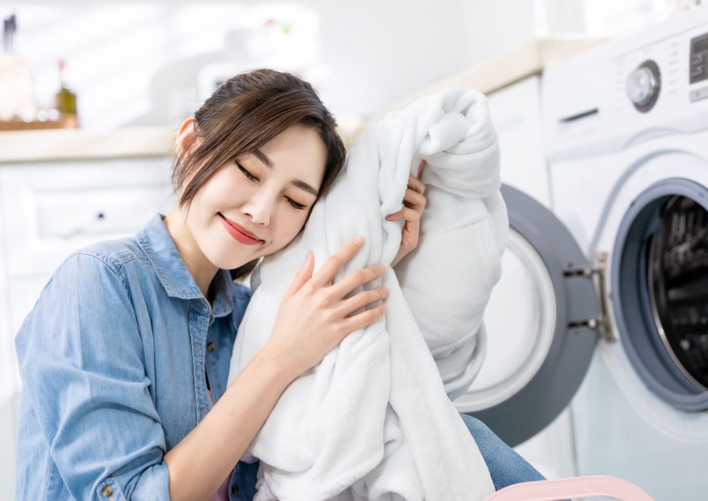 Smiling woman hugging a soft, fresh-smelling white towel beside a front-load washing machine